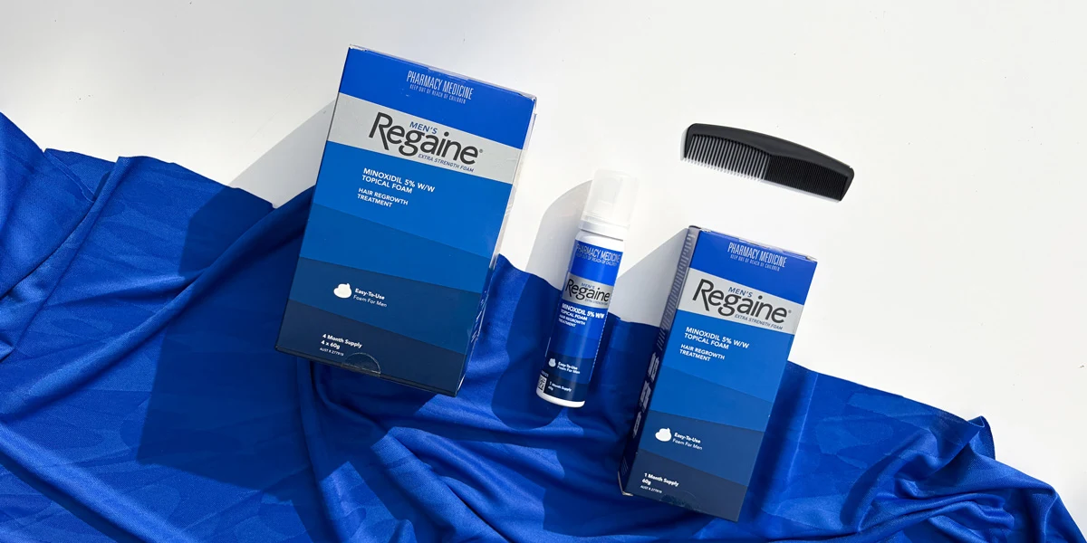 Men's Regaine Minoxidil 5% foam boxes, a canister, and a black comb on a blue fabric and white background.