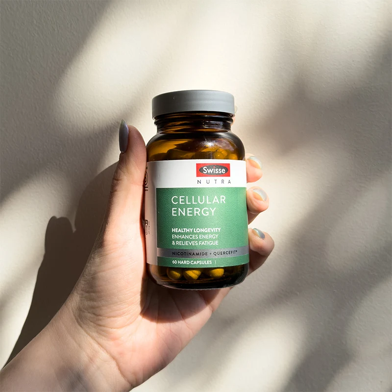 Hand holding a brown glass bottle of Swisse Nutra Cellular Energy supplement with green label against white background with shadows