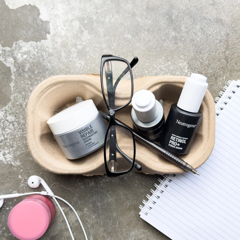 Neutrogena skincare products, glasses, and a pen in a tray beside a notebook, earbuds, and pink container on concrete.