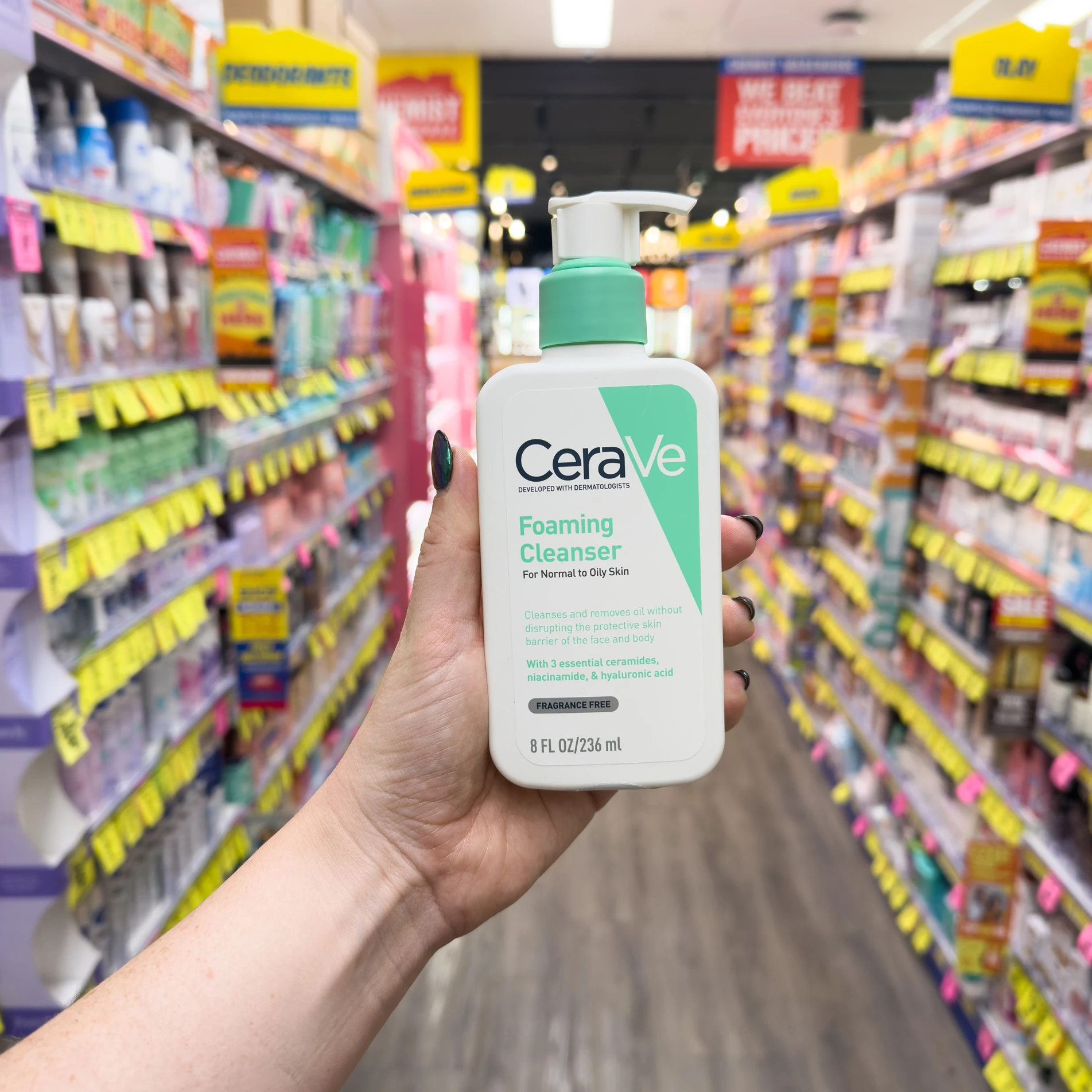 Hand holding CeraVe Foaming Cleanser bottle in a drugstore aisle with colorful product displays and yellow price tags in background