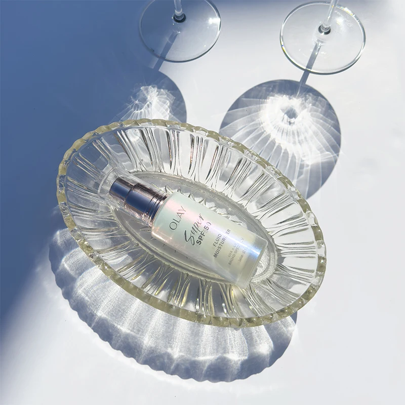 Crystal glass dish with Olay skincare bottle casting dramatic shadows against white background with wine glasses above.