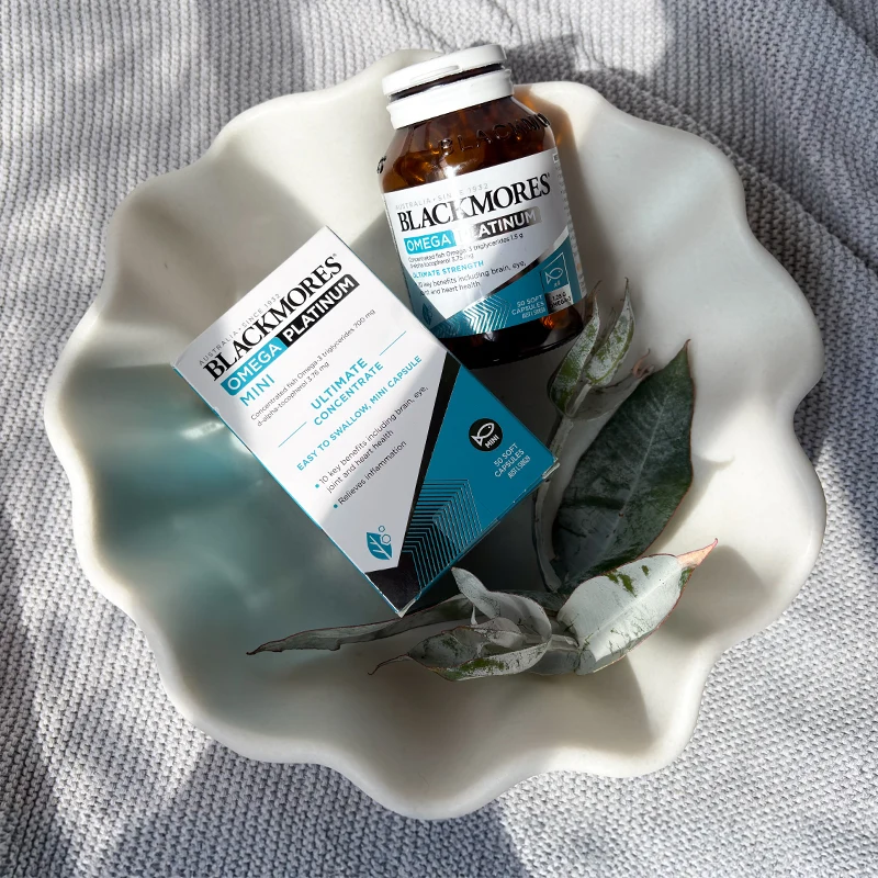 Blackmores Omega Platinum supplements displayed in white scalloped bowl with decorative leaves on grey textured fabric.