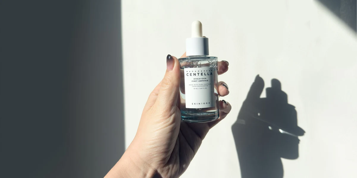 A hand holding a clear glass dropper bottle of SKIN1004 Madagascar Centella Hyalu-Cica First Ampoule against a white wall.