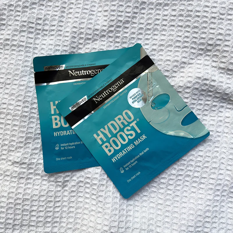 Three teal-colored Neutrogena Hydro Boost hydrating face mask packets arranged on white textured fabric background.