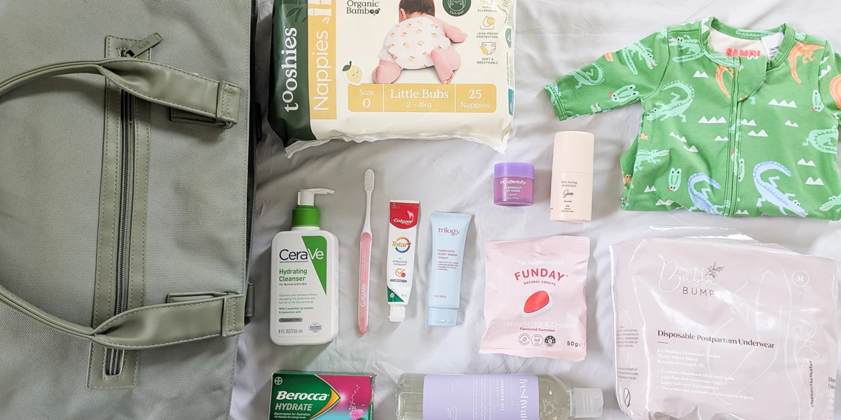 Flat lay of a hospital bag with baby essentials including nappies, CeraVe cleanser, Colgate toothpaste, Trilogy cream, snacks, and a green dinosaur onesie.