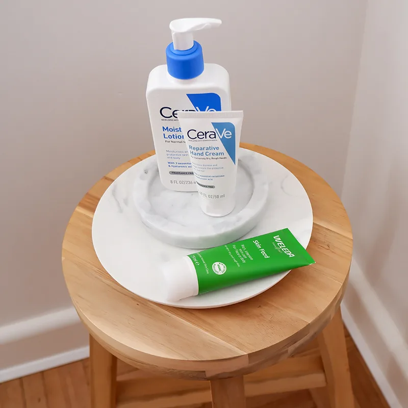 CeraVe skincare products including moisturizing lotion and hand cream displayed on white plate atop wooden stool.
