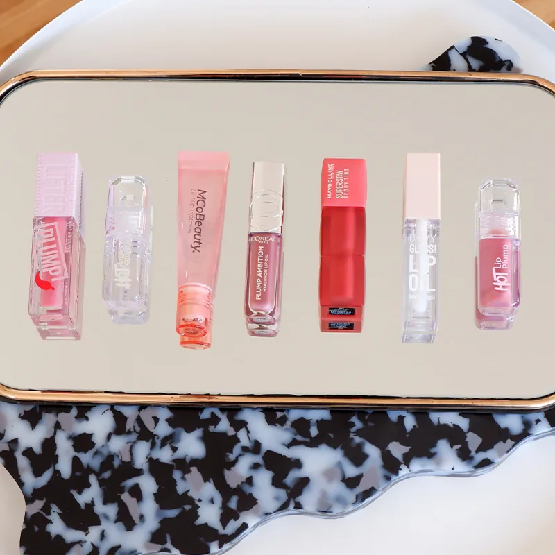 MCoBeauty lip products in pink and nude shades displayed on reflective surface with black and white patterned edge.