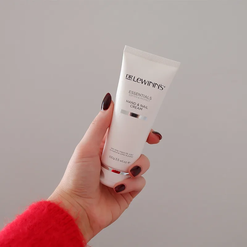Hand with dark manicure holding white tube of Dr Lewinn's hand and nail cream against grey background, wearing red sleeve.