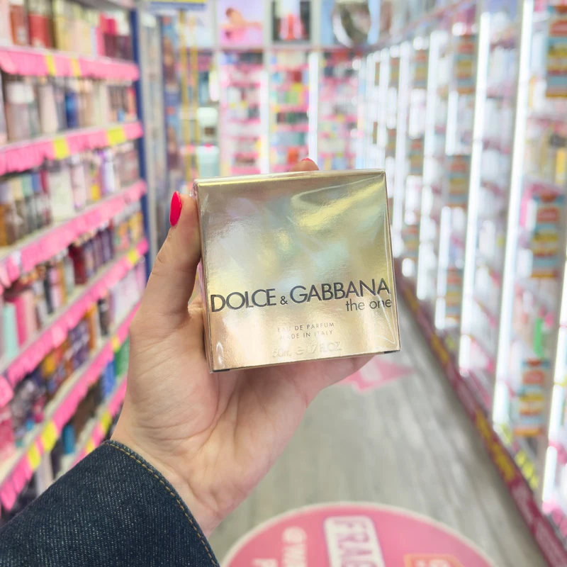 A hand with red nails holds a gold Dolce & Gabbana The One Eau de Parfum box in a fragrance store aisle.