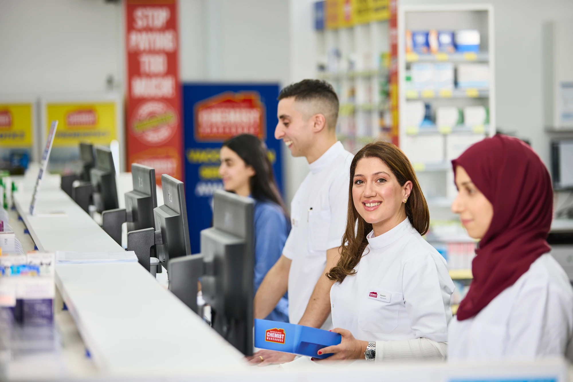 Four Chemist Warehouse staff in white uniforms smile at the pharmacy counter, with shelves of products visible in the background.