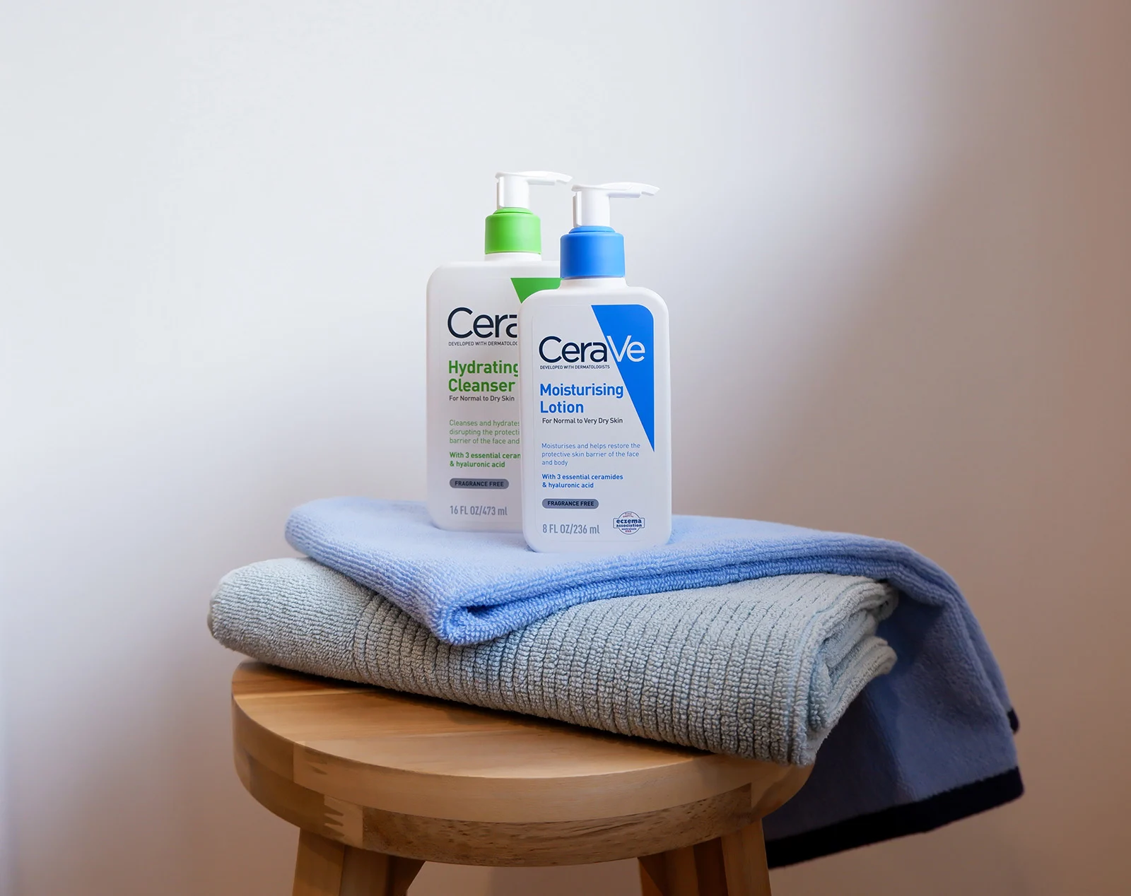 Two CeraVe skincare products, a hydrating cleanser and moisturizing lotion, sitting on folded blue towels on a wooden stool.