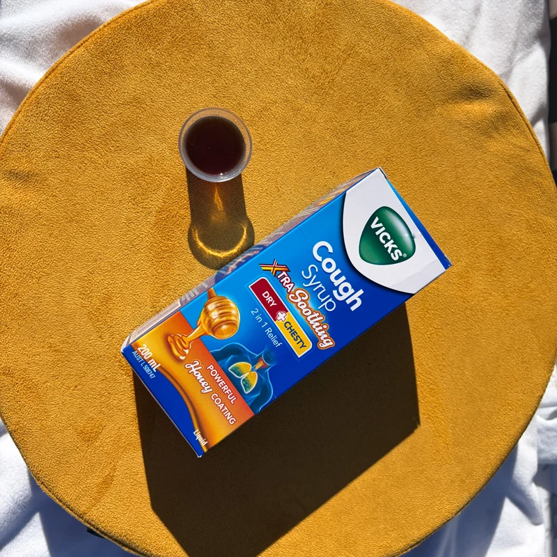 Vicks Cough Syrup Xtra Soothing box and a small medicine cup on a round yellow surface in sunlight.