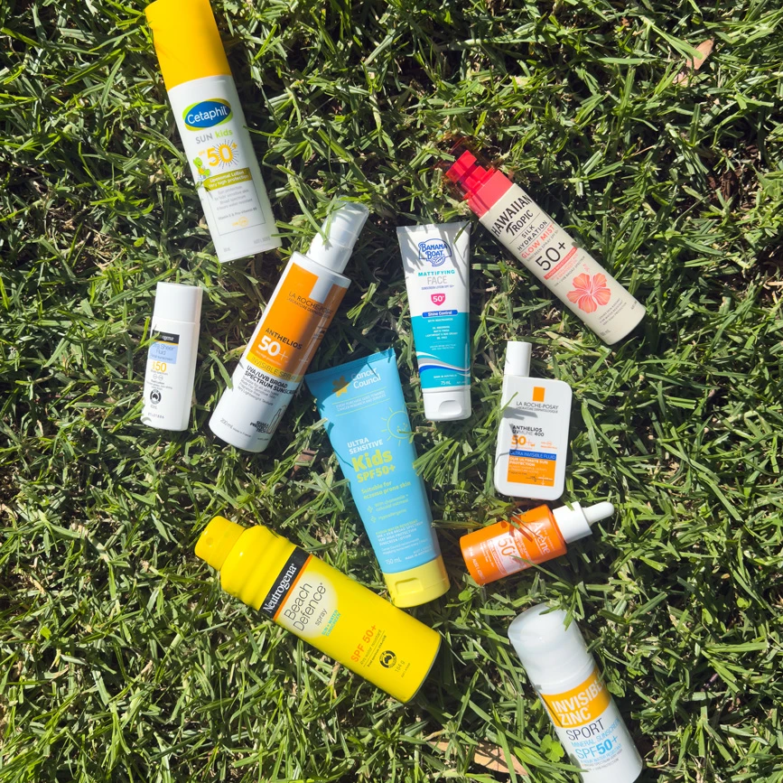 Various sunscreen products from brands like Cetaphil, La Roche-Posay, and Neutrogena scattered on green grass.