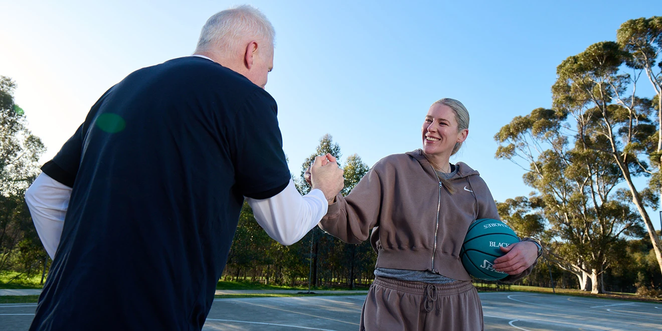 Two people in athletic wear practicing basketball moves outdoors on a sunny day, with eucalyptus trees in the background.