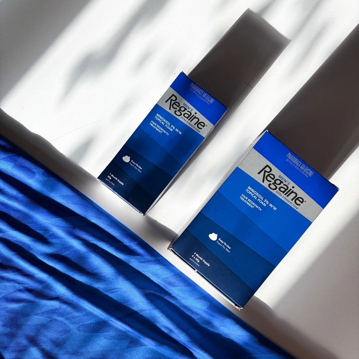 Two blue Regaine Men's Minoxidil 5% hair regrowth foam boxes on a white surface with blue fabric nearby.