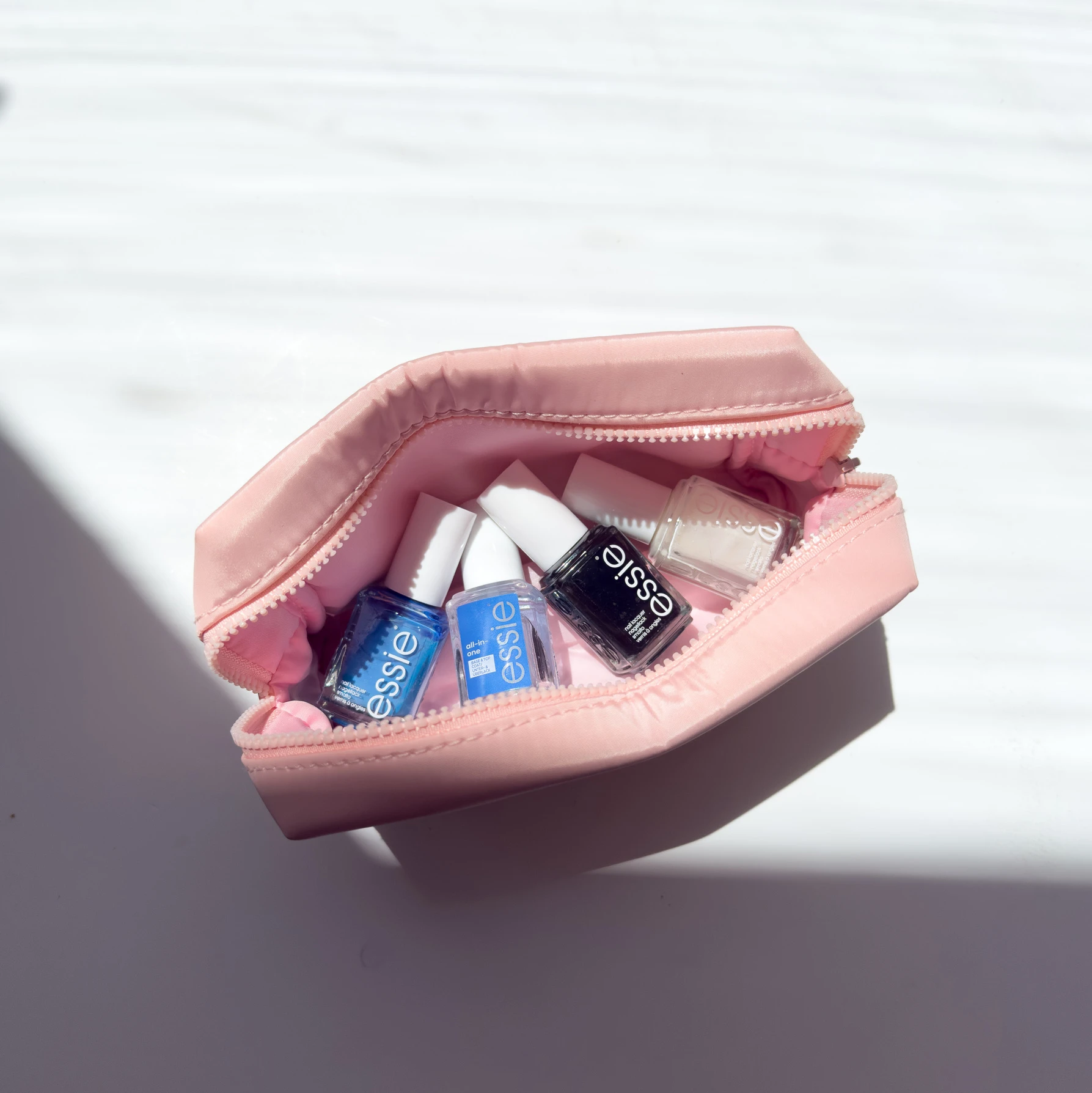 Pink makeup bag unzipped to reveal Essie blue and black nail polish bottles inside, casting a shadow on light surface