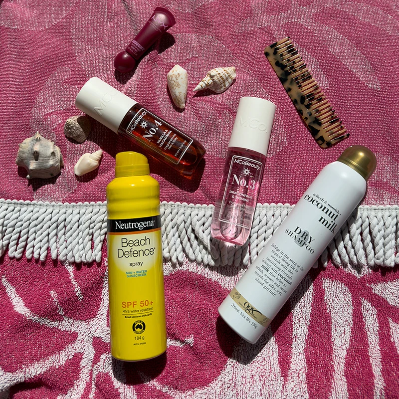 Beauty and sun protection products including sunscreen, nail polish, and hair products arranged with seashells on a pink patterned background.