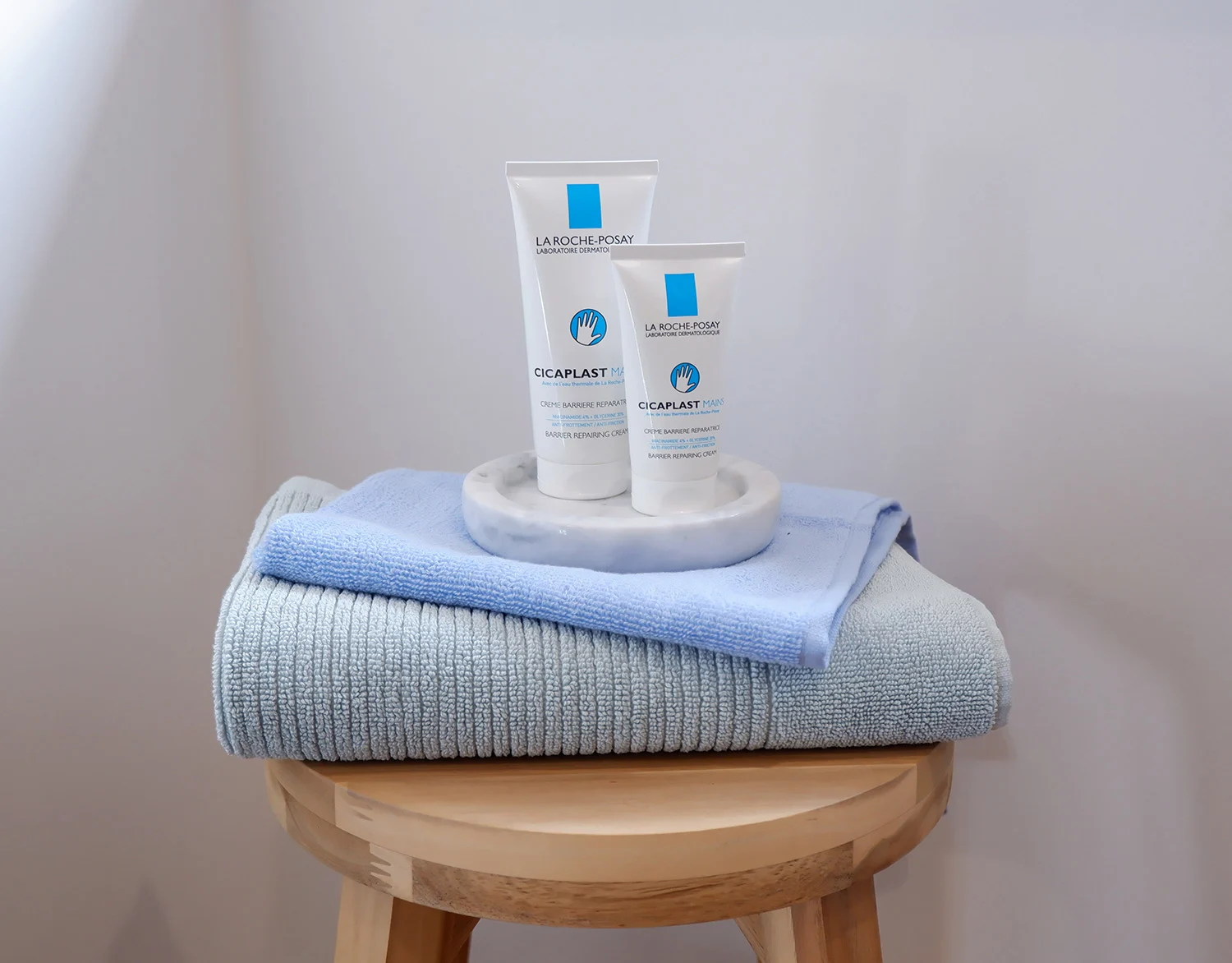 Two La Roche-Posay Cicaplast skincare tubes on marble dish, displayed atop folded blue and gray towels on wooden stool.