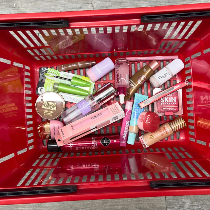 A red shopping basket filled with various makeup products including bronzer, lip gloss, mascara, and skin enhancer, viewed from above.