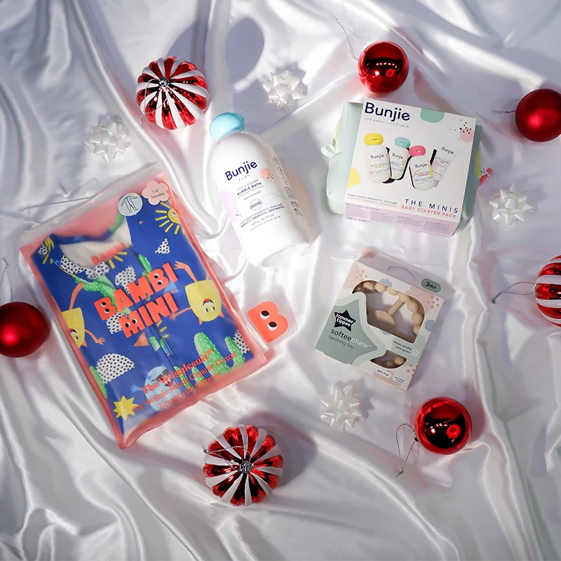 Baby care products and red Christmas ornaments arranged on white satin fabric with a colorful children's book and gift packaging.