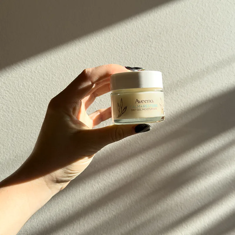 Hand holding Aveeno oat gel moisturizer jar against white wall with dramatic diagonal shadows casting across the scene.