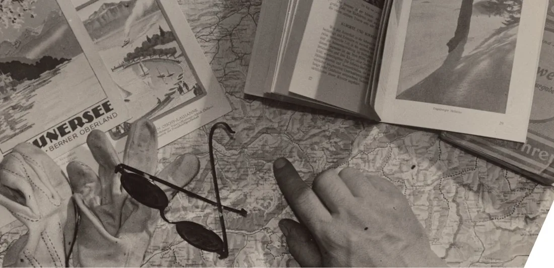 This image shows a travel planning scene for the Swiss Alps, specifically the Berner Oberland region. It includes a map, guidebook, postcards, gloves, sunglasses, and a hand pointing to a spot—likely around Thun or Interlaken.