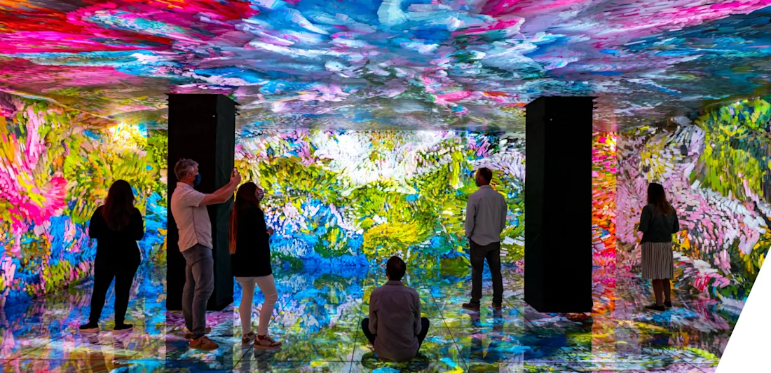 The Gaudi cube at Casa Batlló - six people stand or sit in a space with two dark columns, and walls, ceiling and floor covered in screens showing one huge colourful swirling image