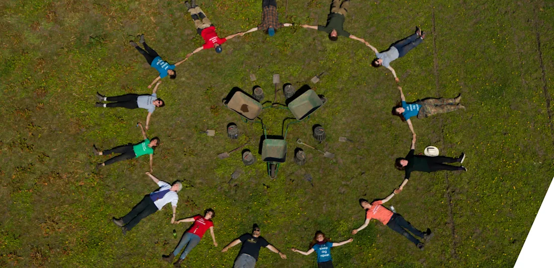 An aerial view of 14 people lying down on grass, hand in hand, forming a circle. In the centre of the circle are archaeology tools - wheel barrows, buckets, spades and trowels