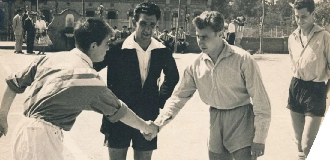 Two football players shake hands in front of a referee. A small crowd of people can be seen in the distance.