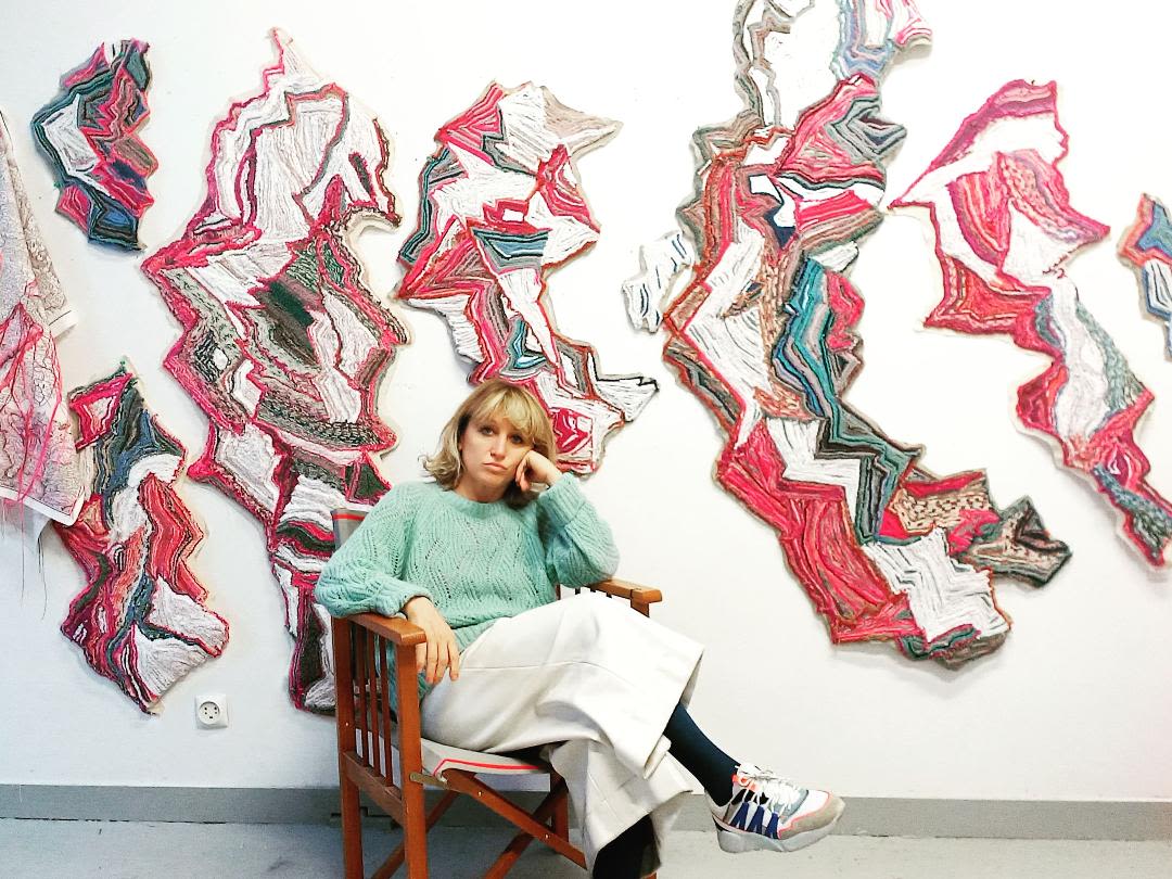 Brankica Zilovic is wearing sneakers, black socks, wide white pants and a teal sweater and lounges casually in a chair. Behind her on the wall is an artwork made by her made up of multicoloured threads woven together into patterns.