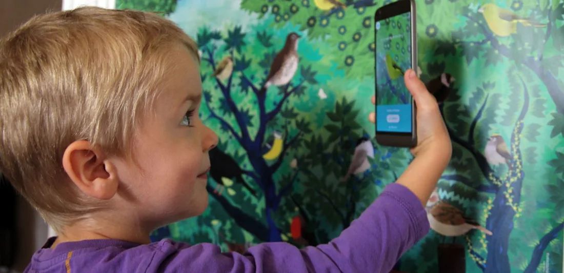 A child holding a phone up to a wall decorated with birds