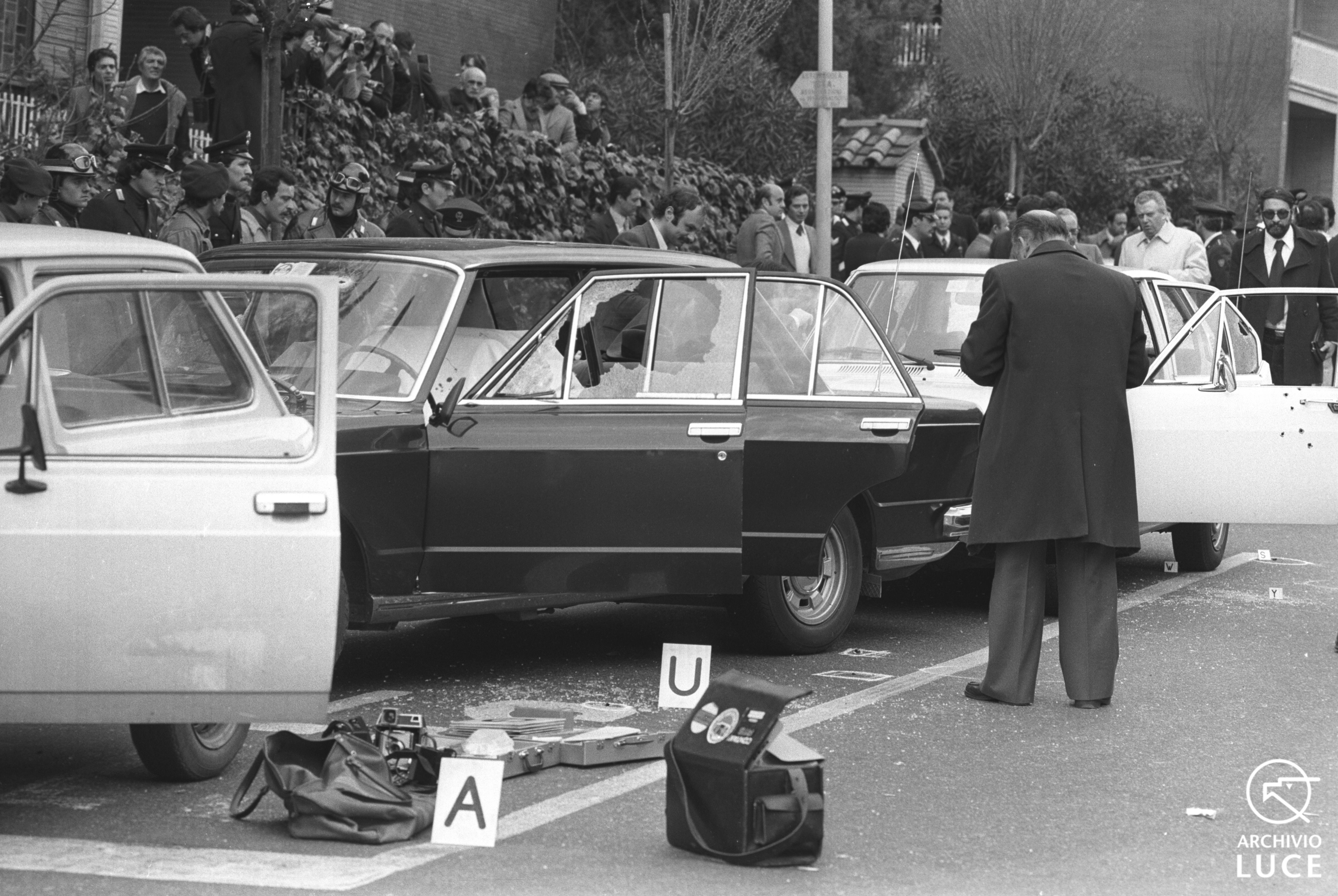 black and white photo of several parked cars, their doors open, their windows smashed and drivers' side doors riddled with bullet holes. A detective stands by to inspect the scene, while onlookers gather in the background.