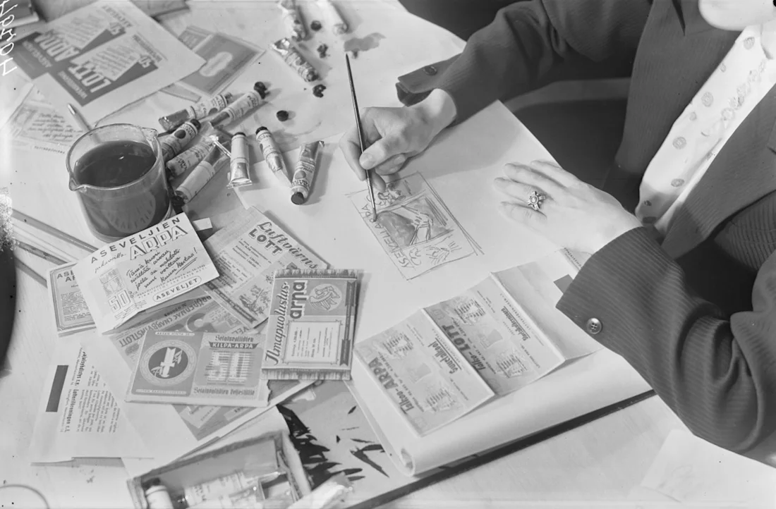 Person painting on paper at a desk covered with ration coupons, spools of thread, and art supplies.