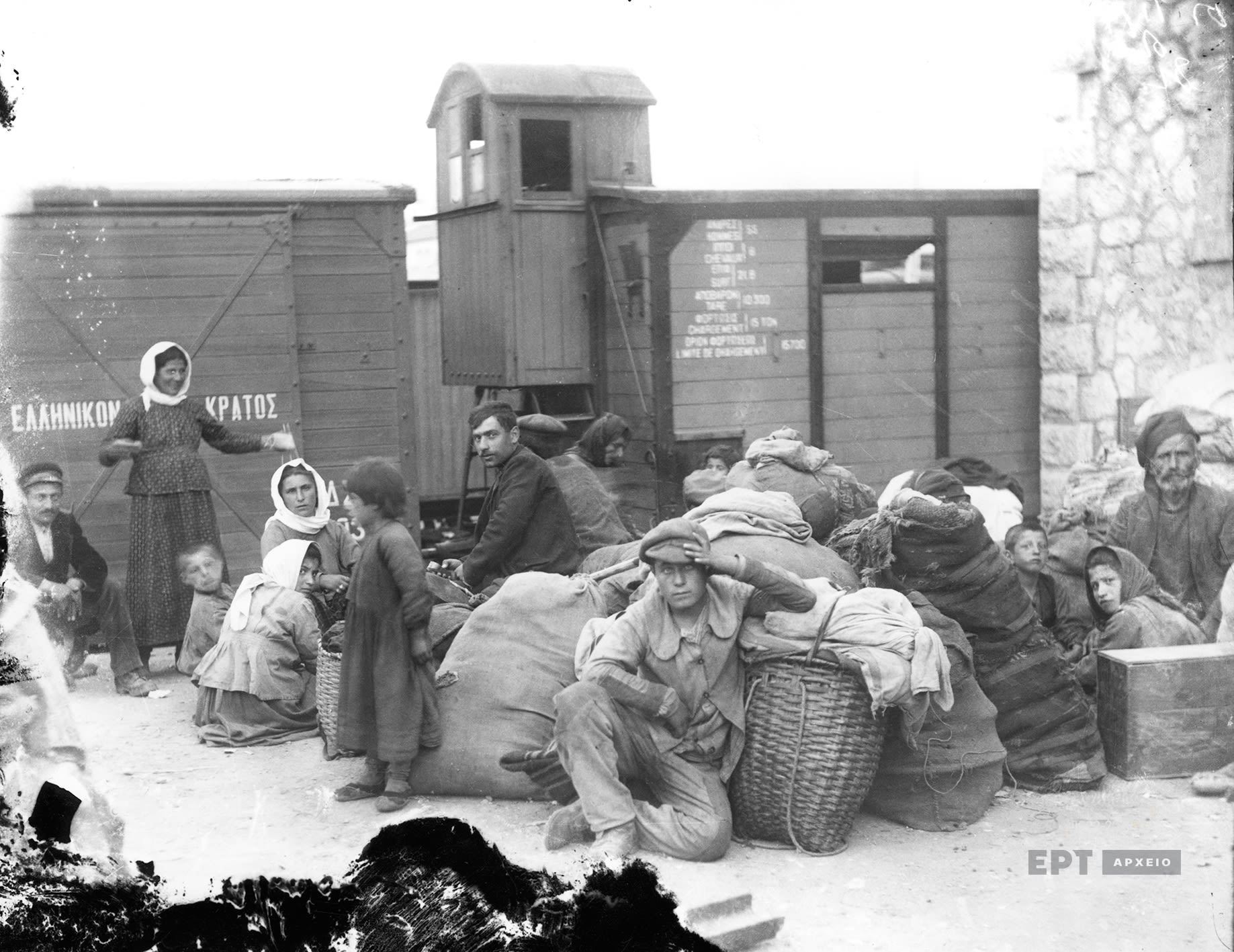 Group of people sitting in front of a train carriage. They have sacks and baskets with their belongings. 