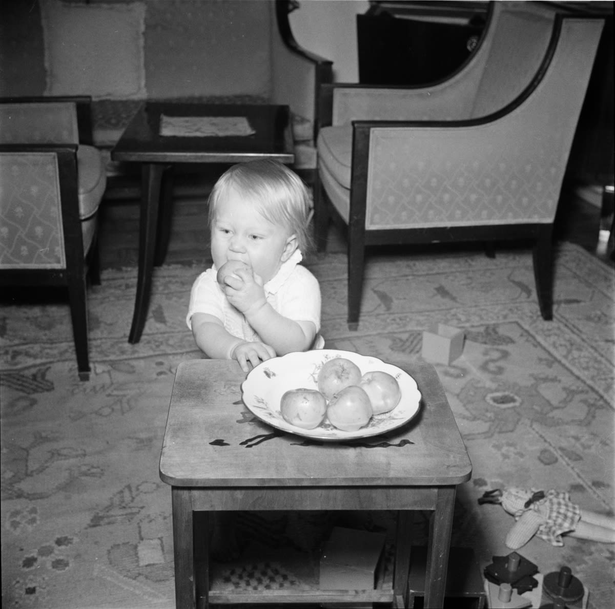 Black and white photograph of a small child biting an apple. A bowl of apples on a low table in front of him. 