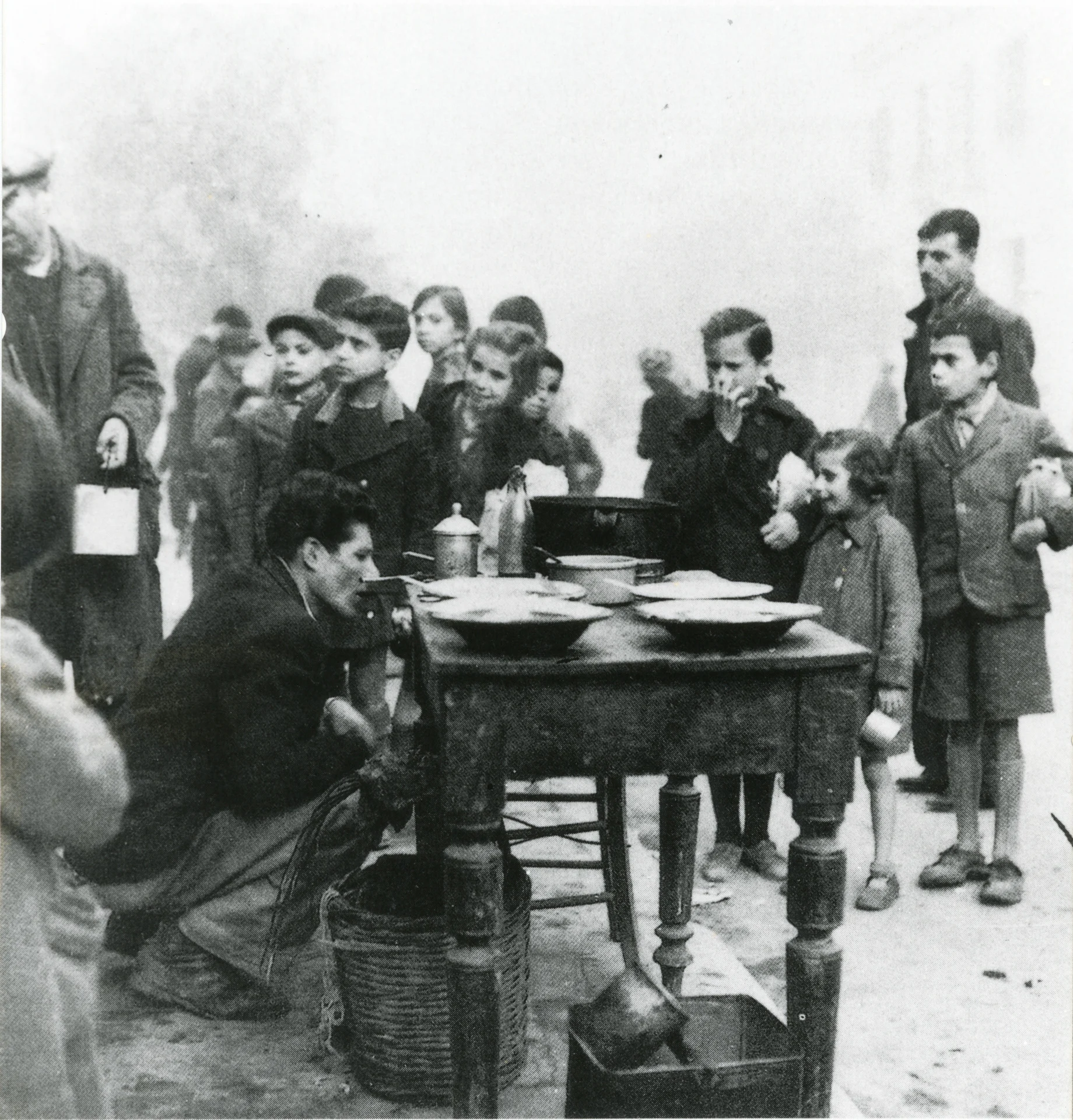 Children gather around a wooden table outdoors, waiting in line for food.