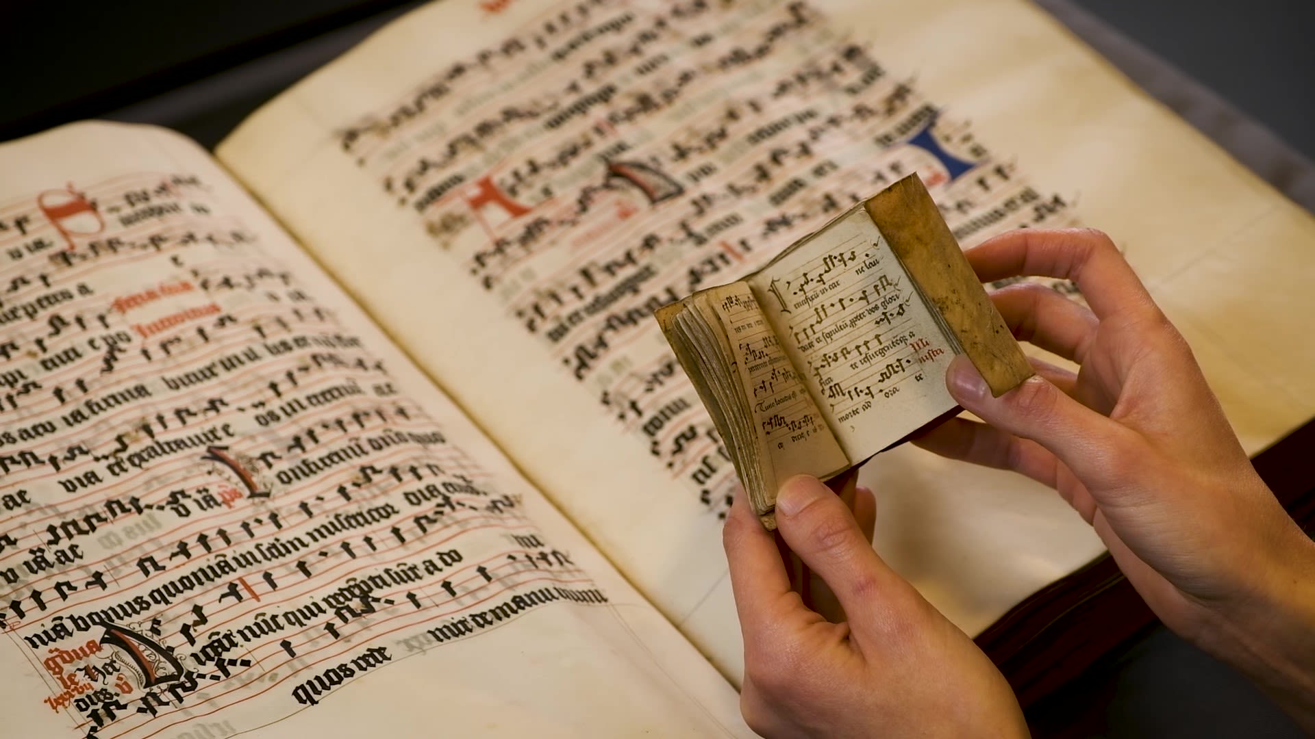 Still frame from a video discussing medieval manuscripts. Two hands holding a very small codex positioned just above a large manuscript book. Both books have detailed calligraphy.