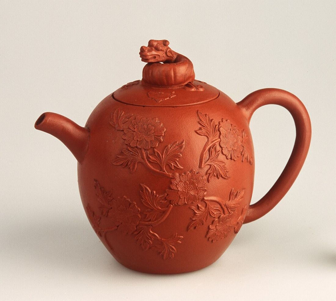 a large clay teapot decorated with clay relief of flowers. The lid has a clay decoration of a dragon on top of it.