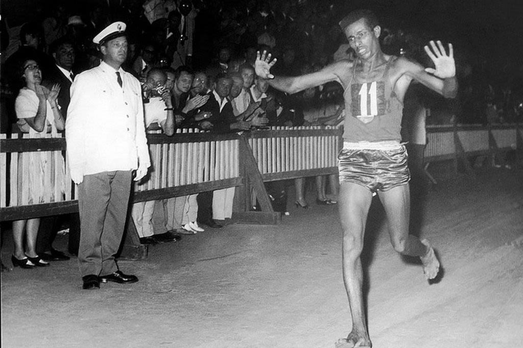  Photographie en noir et blanc d'Abebe Bikila remportant un marathon. Il lève les bras en signe de célébration sous le regard d'un officiel de la course et sous les acclamations de la foule.
