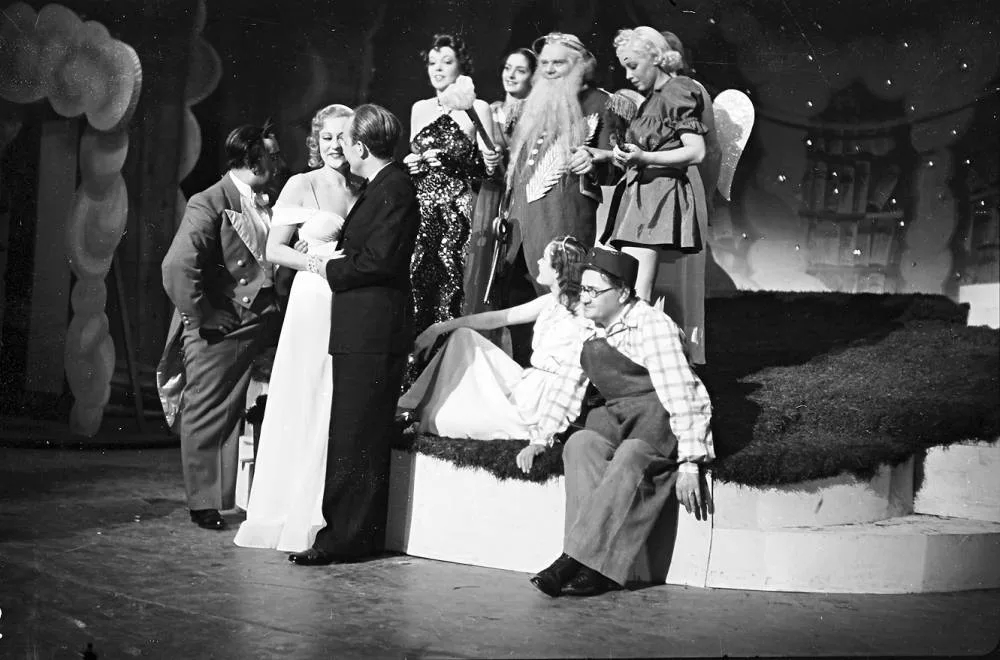 A group of people in costumes gather on a stage set, some standing, others sitting on steps.