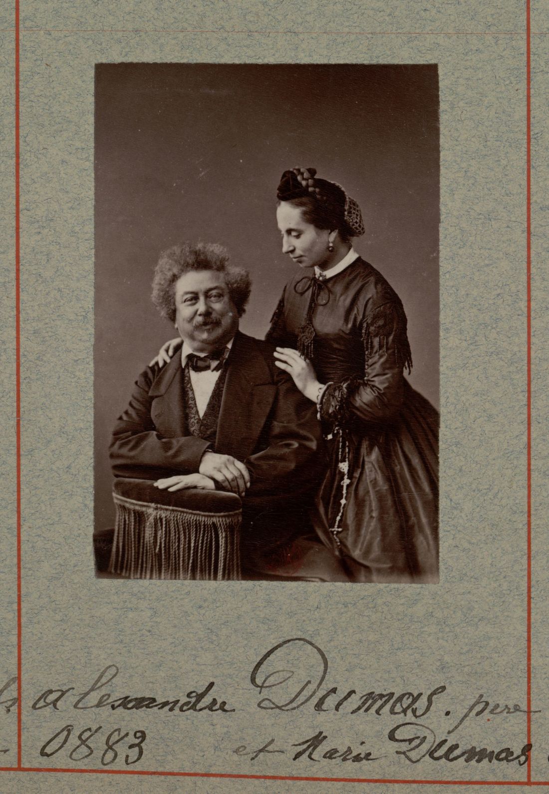Photographie en noir et blanc d'Alexandre Dumas assis, regardant l'appareil photo, tandis que sa fille Marie Dumas est debout et le regarde, les mains sur ses épaules.
