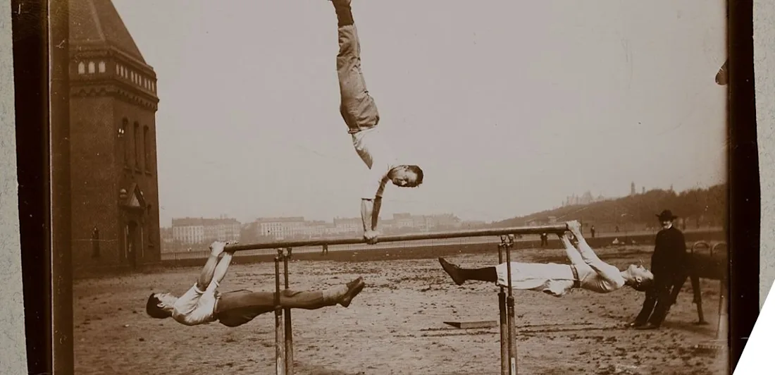 Men balancing on a beam, two holding on underneath each end; one doing a handstand in the middle of the beam. A man in a dark coat and hat looks on.