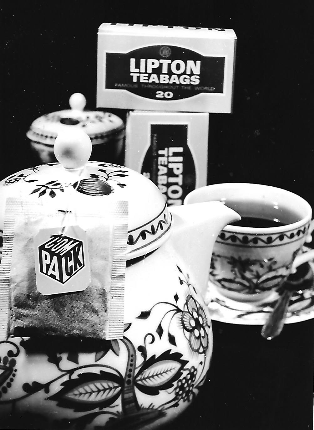 a black and white photograph of a teapot and teacup filled with tea. A tea bag hangs out of the teapot, prominently showing the label which shows the Compack tea company logo. In the background stand boxes of tea bags with the words 'Lipton teabags' on them.