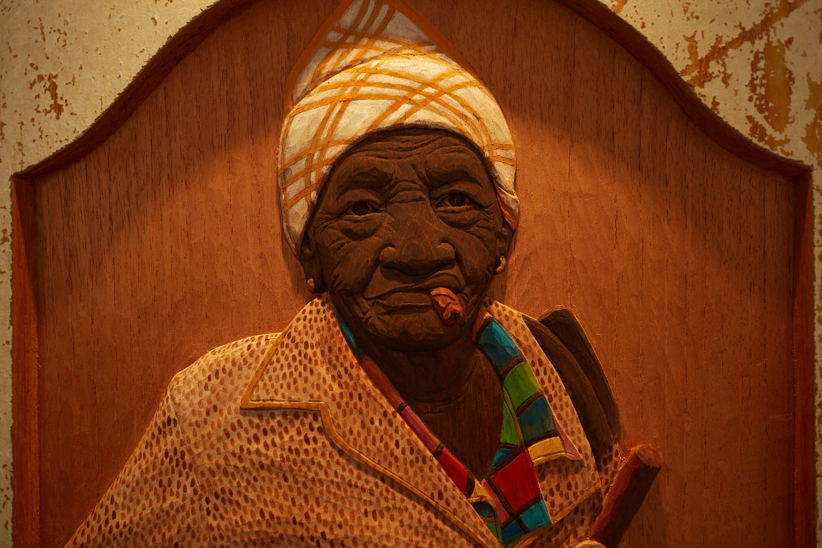 a photograph of a wooden bas-relief on a wall of an old Cuban woman with a cigar in her mouth, wearing a head scarf and a colourful collared shirt.