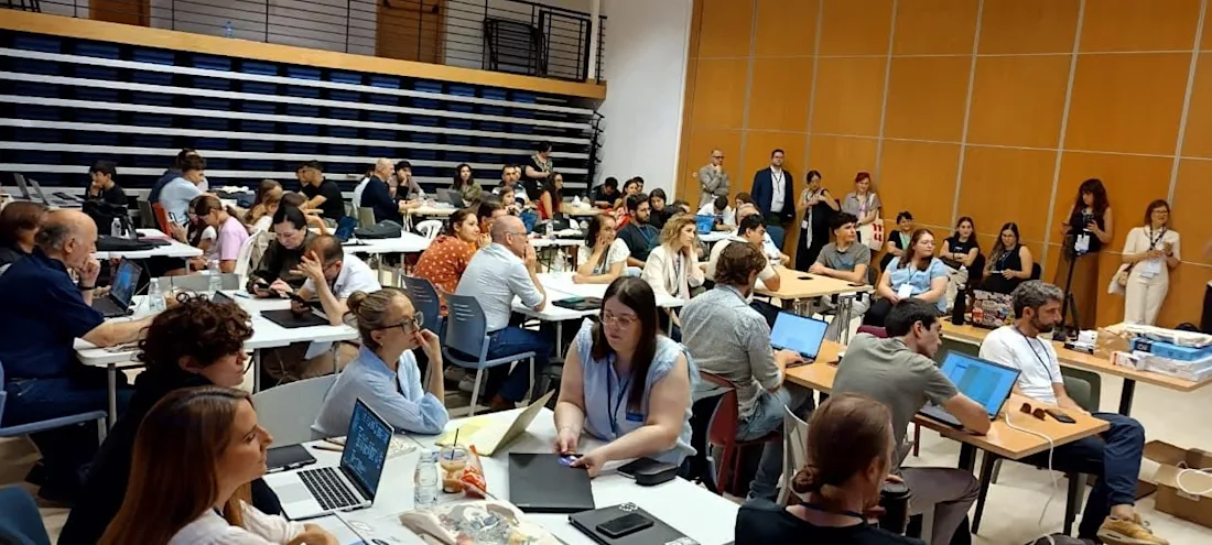 A large group of people sit at tables with laptops in a classroom or workshop setting.