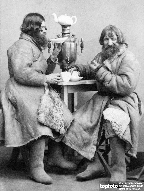 two mean in heavy winter fur clothing sit at a table drinking tea from white porcelain teacups.