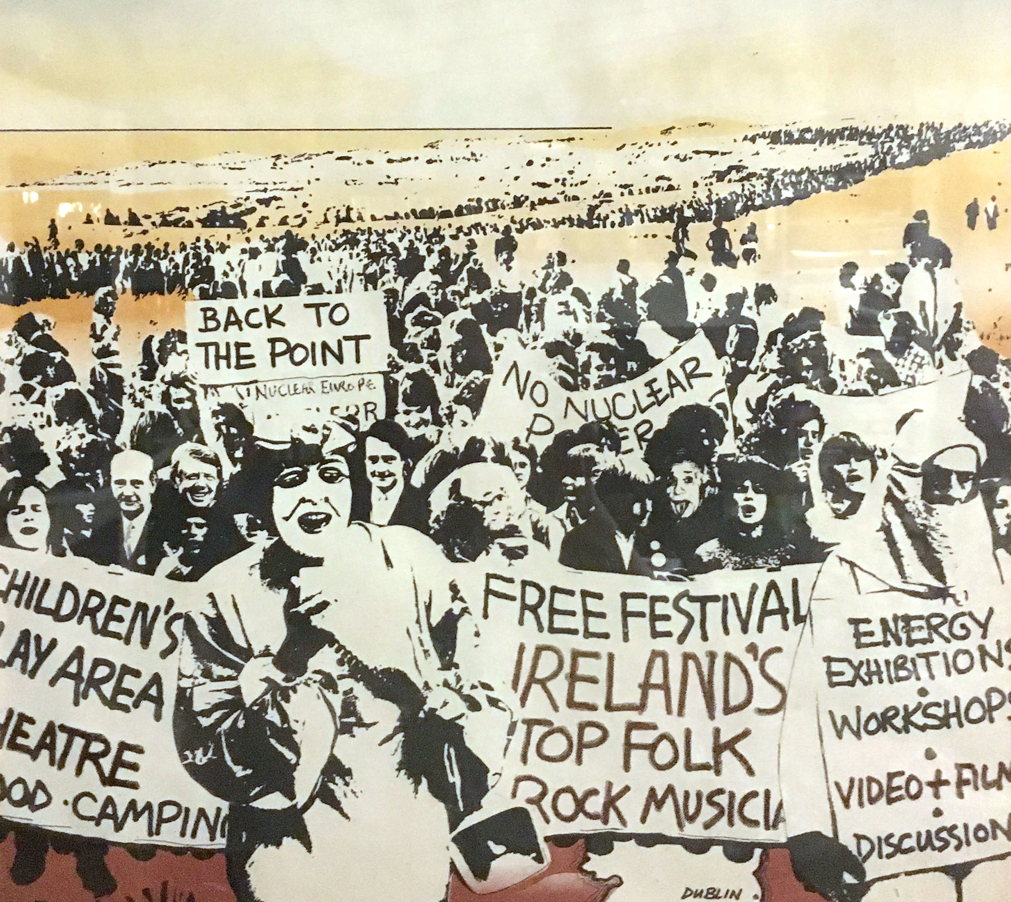 Illustration of a large crowd holding protest and festival signs, including “NO NUCLEAR” and “Ireland’s Top Folk Rock Musical”.