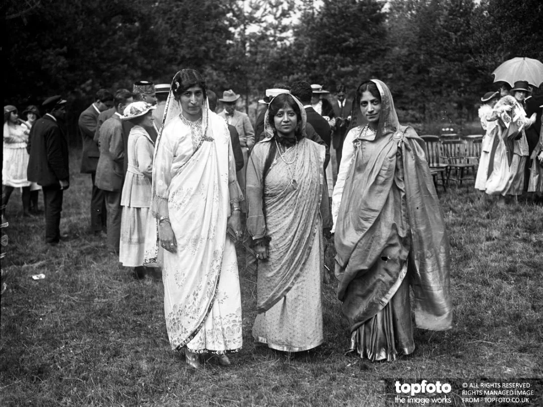 black and white photograph, three women standing in a garden with other people in the background