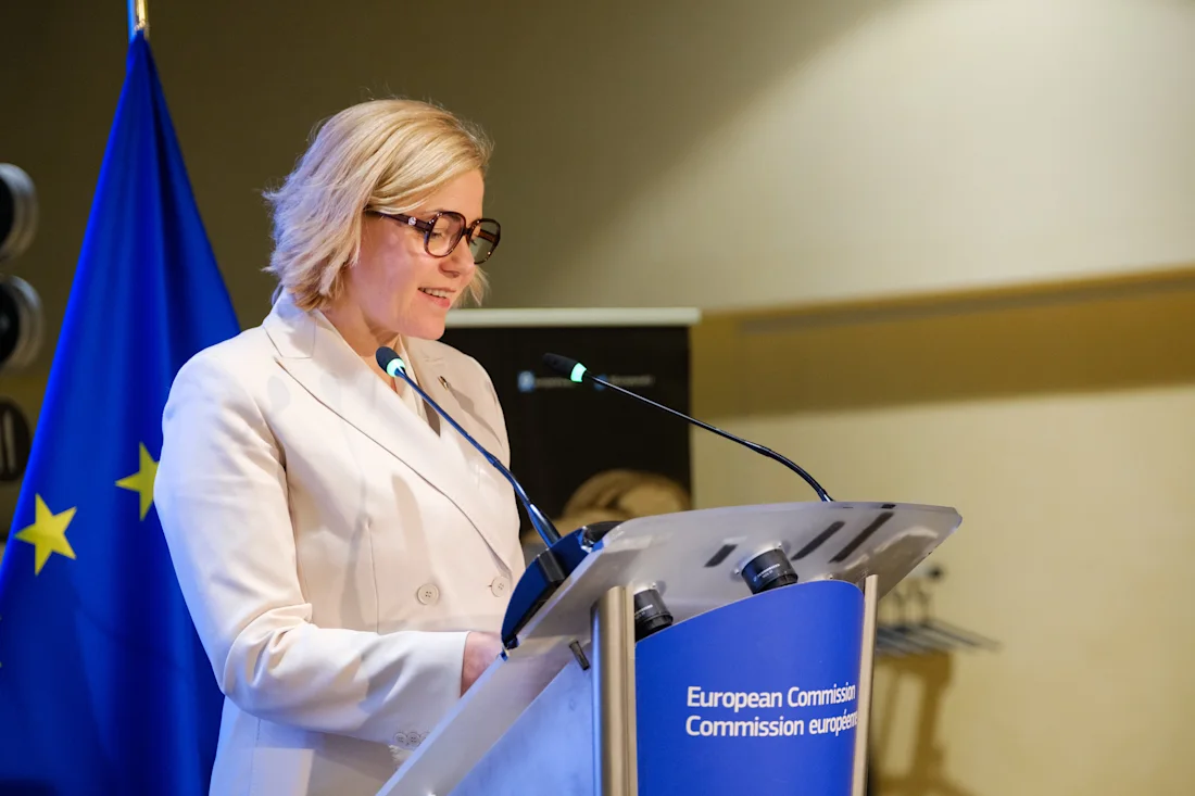 A woman speaks at a podium with a European Commission sign and an EU flag in the background.