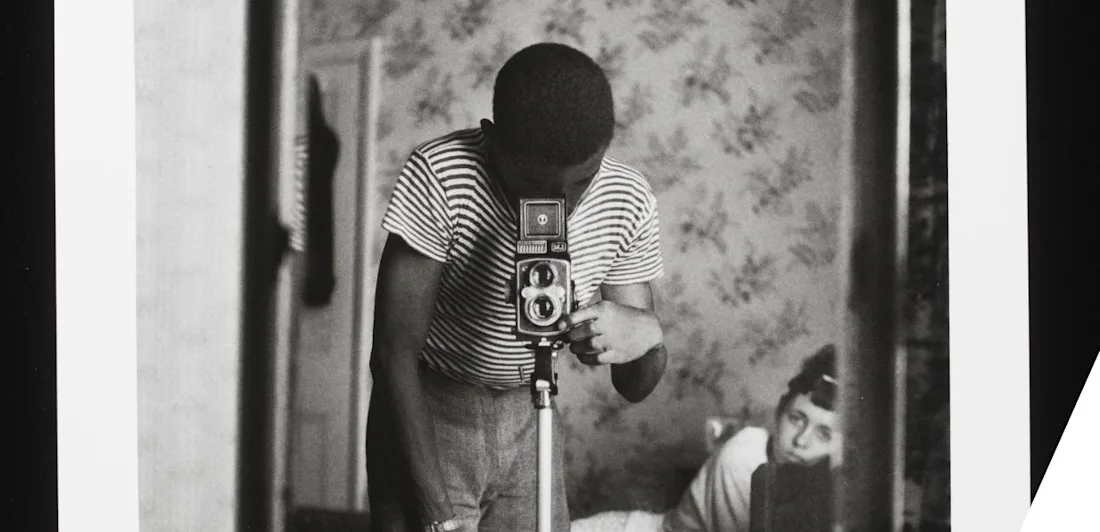 A person taking a picture of themself in a mirror, another looking on from the background